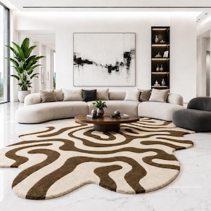 May include: A modern living room with a large, curved beige sofa, a dark gray armchair, and a round wooden coffee table. A unique rug with a brown and beige organic pattern covers the floor. Large windows offer a view of palm trees and a city skyline.