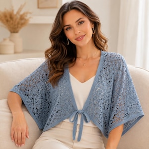 May include: A light blue, open-front, knitted bolero with short sleeves. The bolero features a delicate, openwork design and a tie closure at the waist. The garment is worn over a white top and paired with beige pants.
