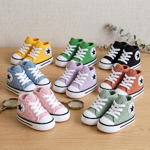 May include: A collection of colorful, crocheted high-top sneakers. The shoes are in various colors, including yellow, green, black, orange, blue, pink, and purple. Each shoe features white laces, a white sole, and a star emblem on the side.