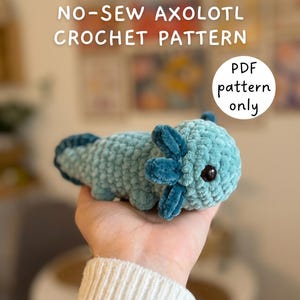 May include: A light blue crocheted axolotl plush toy with dark blue gills and black eyes. The text NO-SEW AXOLOTL CROCHET PATTERN and PDF pattern only is visible in the image. The plush has a soft, textured yarn appearance and a rounded body.