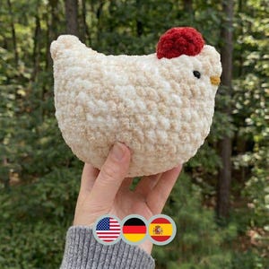 May include: A plush, cream-colored chicken with a red comb and a yellow beak. The handmade stuffed animal is held up against a backdrop of green trees. The chicken has a round body and a small, black eye.