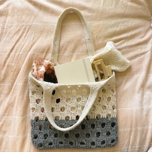 May include: A cream and grey crochet tote bag featuring an openwork pattern, two sturdy handles, and a rectangular base. The bag contains a white notebook, a beige hair claw clip, and a patterned fabric item tucked inside the main compartment.