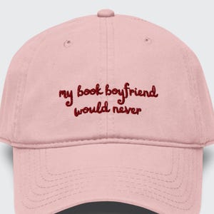 Puede incluir: Una gorra de béisbol de color rosa claro con el texto bordado "my book boyfriend would never" en letras cursivas de color rojo oscuro. La gorra tiene una corona estructurada, una visera curva con costuras y un botón en la parte superior.