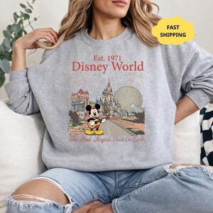 May include: A gray sweatshirt featuring a Disney World graphic. The graphic includes the text "Est. 1971 Disney World" and an illustration of Mickey Mouse playing a guitar in front of various park landmarks. The phrase "The Most Magical Place On Earth" is also present.