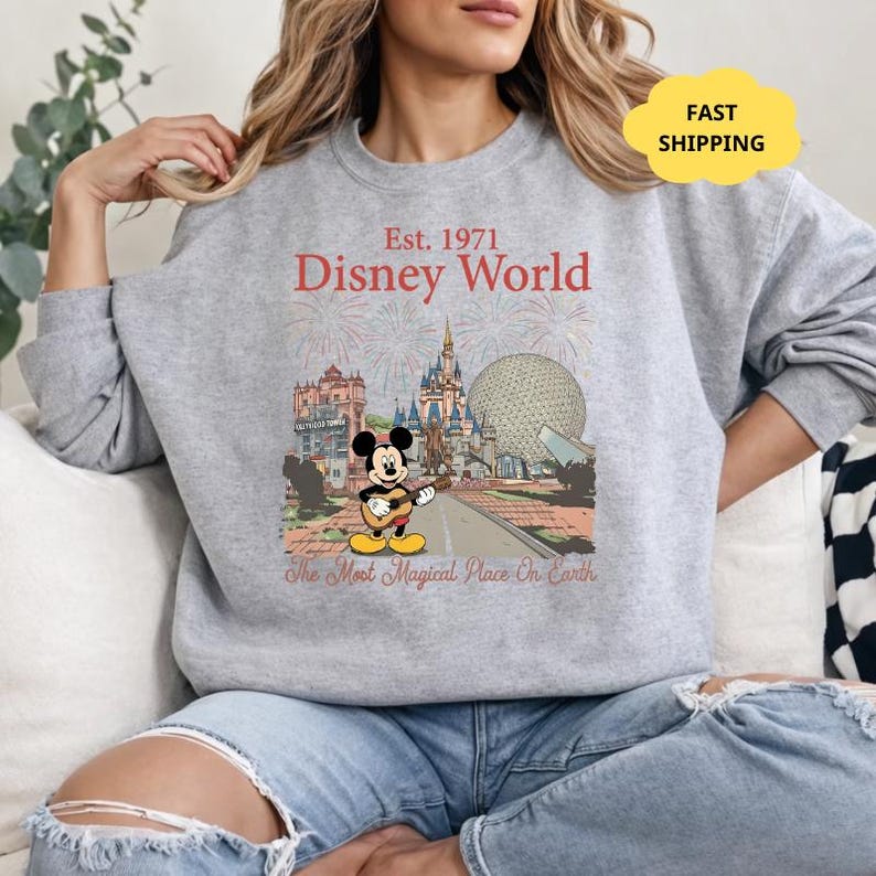 May include: A gray sweatshirt featuring a Disney World graphic. The graphic includes the text "Est. 1971 Disney World" and an illustration of Mickey Mouse playing a guitar in front of various park landmarks. The phrase "The Most Magical Place On Earth" is also present.