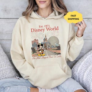 May include: Cream-colored hoodie featuring a Disney World graphic. The design includes the text "Est. 1971 Disney World" with illustrations of iconic park landmarks and Mickey Mouse playing a guitar. The phrase "The Most Magical Place On Earth" is also present.