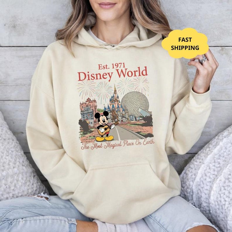 May include: Cream-colored hoodie featuring a Disney World graphic. The design includes the text "Est. 1971 Disney World" with illustrations of iconic park landmarks and Mickey Mouse playing a guitar. The phrase "The Most Magical Place On Earth" is also present.