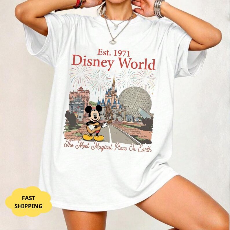 May include: White t-shirt featuring a colorful graphic of Disney World with Mickey Mouse playing a guitar. The text reads "Est. 1971 Disney World" and "The Most Magical Place On Earth".