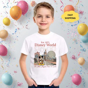 May include: A white t-shirt featuring a graphic of Mickey Mouse playing a guitar in front of Disney World landmarks. The shirt has the text "Est. 1971 Disney World" and "The Most Magical Place On Earth".