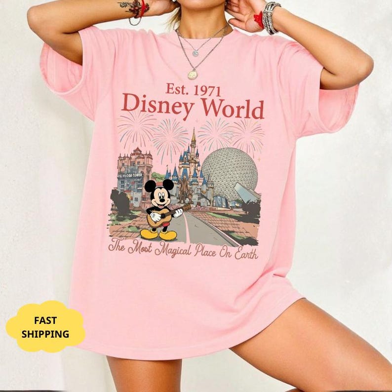 May include: Pink t-shirt featuring a Disney World graphic with Mickey Mouse playing a guitar. The design includes the text "Est. 1971 Disney World" and "The Most Magical Place On Earth". The shirt has a relaxed fit.