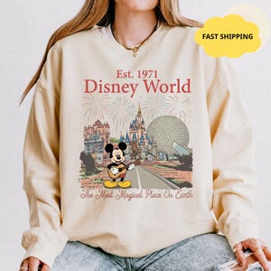 May include: Beige sweatshirt featuring a Disney World graphic. The design includes the text "Est. 1971 Disney World" with illustrations of fireworks, a castle, and Mickey Mouse playing a guitar. The phrase "The Most Magical Place On Earth" is also included.