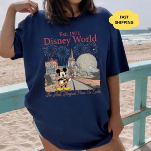 May include: Navy blue t-shirt featuring a Disney World graphic. The design includes the text "Est. 1971 Disney World" with fireworks, Mickey Mouse playing a guitar, and iconic park landmarks. The phrase "The Most Magical Place On Earth" is also included.