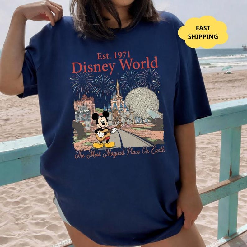 May include: Navy blue t-shirt featuring a Disney World graphic. The design includes the text "Est. 1971 Disney World" with fireworks, Mickey Mouse playing a guitar, and iconic park landmarks. The phrase "The Most Magical Place On Earth" is also included.