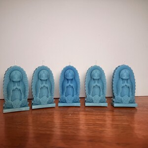 Puede incluir: Cinco velas de la Virgen María de color azul claro. Cada vela es una figura detallada de la Virgen María, rodeada por un halo y rosas en la base. Las velas están sobre una superficie de madera sobre un fondo blanco.