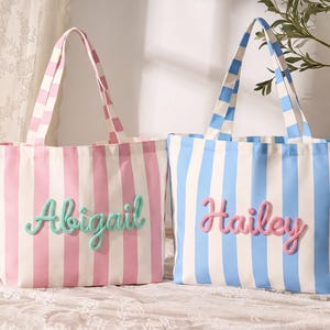 Könnte beinhalten: Zwei Tragetaschen aus Segeltuch mit vertikalen Streifen. Die linke Tasche ist rosa-weiß mit dem Namen Abigail in erhabener mintgrüner Schreibschrift. Die rechte Tasche ist blau-weiß mit dem Namen Hailey in erhabener rosa Schreibschrift gestaltet.