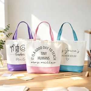 May include: Three canvas tote bags with coloured bases and handles. One has a purple base and handle, another pink, and the third blue. Each bag has text: "teacher Mrs G.", "IT IS A GOOD DAY TO TEACH TINY HUMANS mrs. miller", and "s. Garcia's TEACHER STUFF".