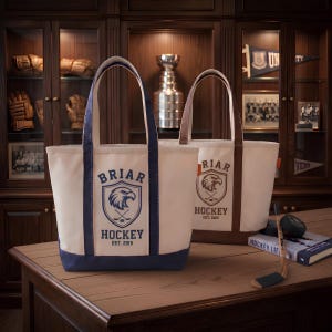 May include: Two canvas tote bags featuring a screen-printed logo with an eagle head inside a shield, crossed hockey sticks, and the text BRIAR HOCKEY EST. 2015. One bag has navy blue trim and base, while the other has brown trim and base.