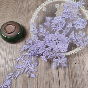 May include: A lavender floral lace applique featuring intricate embroidered flower and leaf patterns on a sheer mesh base. The lace is draped over a circular embroidery hoop decorated with small white pearls, next to a small green ceramic bowl.