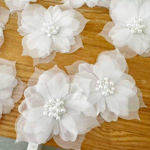 Può includere: Una collezione di fiori in tessuto bianco con petali stratificati e traslucidi, caratterizzati da un grappolo centrale di piccole perle artificiali bianche e rotonde. Ogni fiore è realizzato in materiale delicato e trasparente.