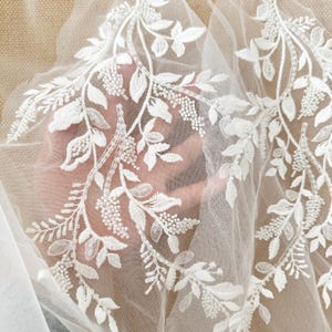 May include: A sheer white tulle fabric featuring intricate white embroidery in a botanical leaf and vine pattern. The design is embellished with small white beads and clear sequins that catch the light, creating a textured and dimensional appearance.