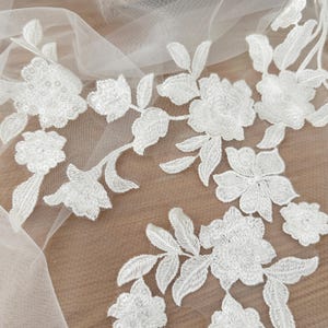 May include: White floral lace applique featuring intricate embroidered flowers and leaves on a sheer tulle mesh base. The design includes delicate stitching and subtle sequin accents throughout the petals, creating a continuous botanical pattern.