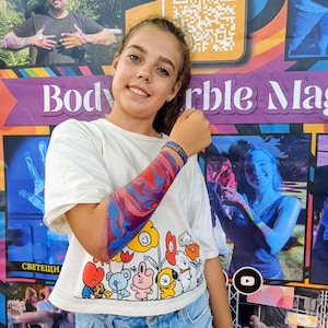 Può includere: Una t-shirt bianca a maniche corte con una stampa grafica colorata di personaggi dei cartoni animati e fiori sul davanti. La maglietta è abbinata a jeans azzurri. Un braccio mostra un vivace design di pittura marmorizzata multicolore.
