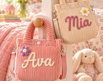 Peut inclure: Deux sacs à main en velours côtelé avec poignées supérieures et bandoulières amovibles. Le sac rose porte le nom Ava en laine crème et une fleur violette. Le sac beige porte le nom Mia en laine rose et une fleur blanche. Fermetures à pression.