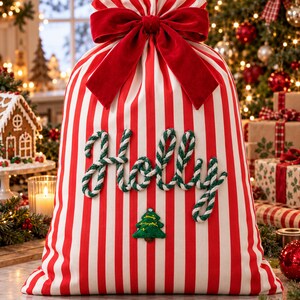 Può includere: Un sacco regalo in tessuto a righe verticali rosse e bianche, chiuso in alto da un grande fiocco in velluto rosso. La parola Holly è scritta in corda intrecciata verde e bianca, con un piccolo albero di Natale ricamato sotto.