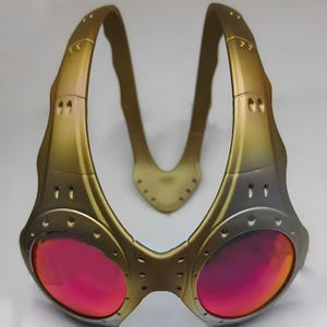 Authentische goldene Oakley Over-The-Top (OTT) Sonnenbrille auf weißem Grund. Das seltene Archive-Piece besticht durch einen matten Goldrahmen in skulpturalem Over-the-head Design. Die kreisrunden Spiegelgläser zeigen einen Verlauf von leuchtendem Gelbgold zu Orange. Mit industriellen Belüftungsschlitzen und Nieten-Details ist dieses Sammlerstück ein Highlight für Techwear-, Gorpcore- und Avantgarde-Enthusiasten sowie Oakley-Liebhaber.