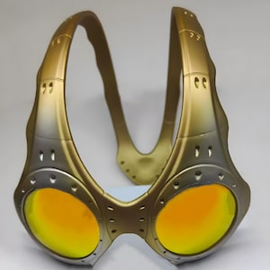 Authentische goldene Oakley Over-The-Top (OTT) Sonnenbrille auf weißem Grund. Das seltene Archive-Piece besticht durch einen matten Goldrahmen in skulpturalem Over-the-head Design. Die kreisrunden Spiegelgläser zeigen einen Verlauf von leuchtendem Gelbgold zu Orange. Mit industriellen Belüftungsschlitzen und Nieten-Details ist dieses Sammlerstück ein Highlight für Techwear-, Gorpcore- und Avantgarde-Enthusiasten sowie Oakley-Liebhaber.