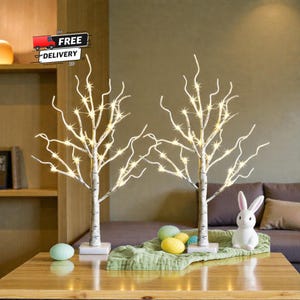 Puede incluir: Dos árboles decorativos de abedul con luces LED blancas cálidas. Los árboles están sobre bases blancas y forman parte de una exhibición de Pascua. Un conejito blanco y huevos coloridos también están en exhibición.