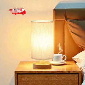 May include: A cylindrical table lamp with a beige fabric shade and a wooden base sits on a wooden nightstand. A white teacup and saucer are next to the lamp. The words "FREE DELIVERY" are in the upper left corner.