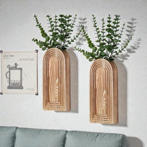 Op de afbeelding: Twee houten wandplantenbakken met een boogvormig ontwerp, elk met groene bladplanten. De plantenbakken zijn gemaakt van lichtgekleurd hout en zijn aan een witte muur bevestigd. Een ingelijste vintage print staat ook op de muur.