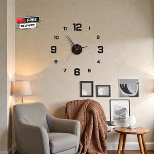 Op de afbeelding: Een moderne wandklok met zwarte cijfers en wijzers tegen een neutrale muur. De wijzerplaat van de klok heeft een minimalistisch ontwerp, met de cijfers 1 tot en met 12. De klok maakt deel uit van een woonkamersetting met een stoel, een bijzettafel en kunstwerken.