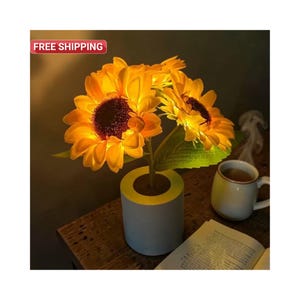 May include: A decorative arrangement of illuminated sunflowers in a light blue and yellow pot. The sunflowers have bright yellow petals and dark brown centers. A white mug and open book are on a wooden surface.