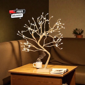 Op de afbeelding: Een decoratieve boomvormige lamp met warmwitte LED-verlichting. De goudkleurige takken worden verlicht door kleine lampjes, waardoor een zachte gloed ontstaat. De lamp staat op een vierkante voet op een houten tafel, naast een kop koffie en een open boek.