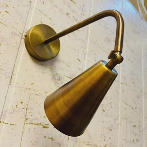 以下が含まれることがあります： 真鍮色の壁掛けランプ。円形のベース、湾曲したアーム、円錐形のシェードを備え、温かみのあるメタリック仕上げです。モダンでミニマリストなデザインです。