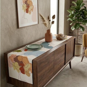 Puede incluir: Un camino de mesa rectangular largo con fondo blanco y dibujos lineales abstractos de árboles y formas orgánicas en tonos naranja, amarillo, marrón y azul. El camino está colocado sobre un aparador de madera oscura con un jarrón y un plato.