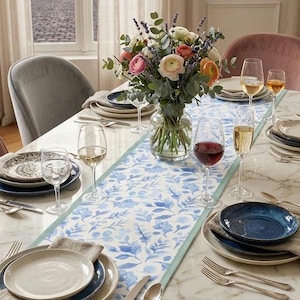 May include: A long rectangular table runner featuring a blue and white watercolour leaf pattern with a solid light blue border. The runner is centred on a white marble dining table set with blue and white plates, wine glasses, and silverware.