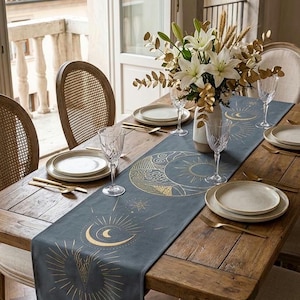 May include: A long, dark gray table runner featuring gold-colored celestial prints of suns, moons, and stars. The runner is laid across a rustic wooden dining table set with white plates, clear crystal glasses, gold-toned cutlery, and a floral centerpiece.