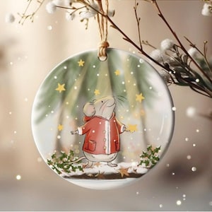 以下が含まれることがあります： 緑と白の冬のシーンが描かれた丸い白いクリスマスオーナメント。赤いコートを着た小さな茶色のネズミが雪の中に立って、星を見上げています。
