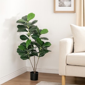 May include: A faux fiddle leaf fig tree with large, dark green leaves and a black pot. The artificial plant is positioned in a corner next to a beige sofa and a framed picture.