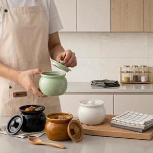 May include: A collection of round ceramic storage jars with matching lids in sage green, glossy black, mustard yellow, and white. The jars feature a wide opening and a smooth finish, suitable for food storage, alongside a wooden spoon.
