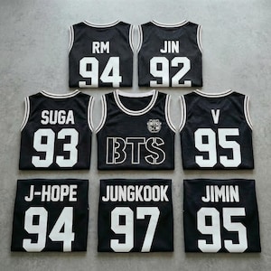 Op de afbeelding: Zwarte basketbalshirts met witte randen en letters. Elk shirt toont een naam en nummer, waaronder "RM 94", "JIN 92", "SUGA 93", "V 95", "J-HOPE 94", "JUNGKOOK 97" en "JIMIN 95". Eén shirt toont "BTS" in grote witte letters.
