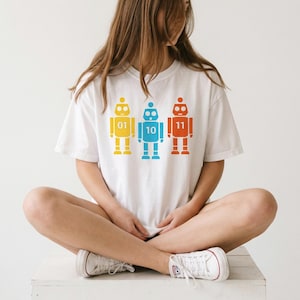 May include: White t-shirt featuring three colorful robot designs. Each robot is a different color: yellow, blue, and red, with the numbers 01, 10, and 11 displayed on their chests. The shirt is a casual, unisex style.
