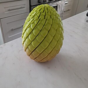 Pu&ograve; includere: Un oggetto a forma di uovo color verde lime con una superficie ruvida simile a delle scaglie. La struttura presenta distinti strati o segmenti orizzontali che avvolgono l'intera forma, creando un aspetto stratificato dalla cima alla base.
