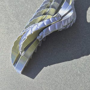 Pu&ograve; includere: Un oggetto stampato in 3D con una forma curva simile a un'ala, caratterizzato da una serie di segmenti impilati e stratificati. La struttura presenta una finitura metallica iridescente che varia tra tonalit&agrave; di blu, viola e argento, con linee di stampa orizzontali visibili.