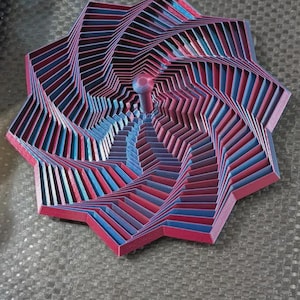 May include: A multi-layered, geometric sculpture with a star-like shape. The artwork features alternating layers of red and blue, creating a spiral effect. A small, rounded, pinkish-red sphere is at the center. The piece is on a gray surface.