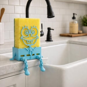 May include: A yellow sponge with a cartoon face sits in a light blue holder with legs, placed on a white countertop near a kitchen sink. The sponge holder has a tie design. A black faucet is visible in the background.