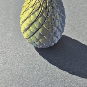 Pu&ograve; includere: Un oggetto a forma di uovo con una superficie strutturata simile a scaglie. L'oggetto presenta una sfumatura di colore che passa da un giallo brillante nella parte superiore a un grigio scuro e spento nella parte inferiore, con creste verticali.